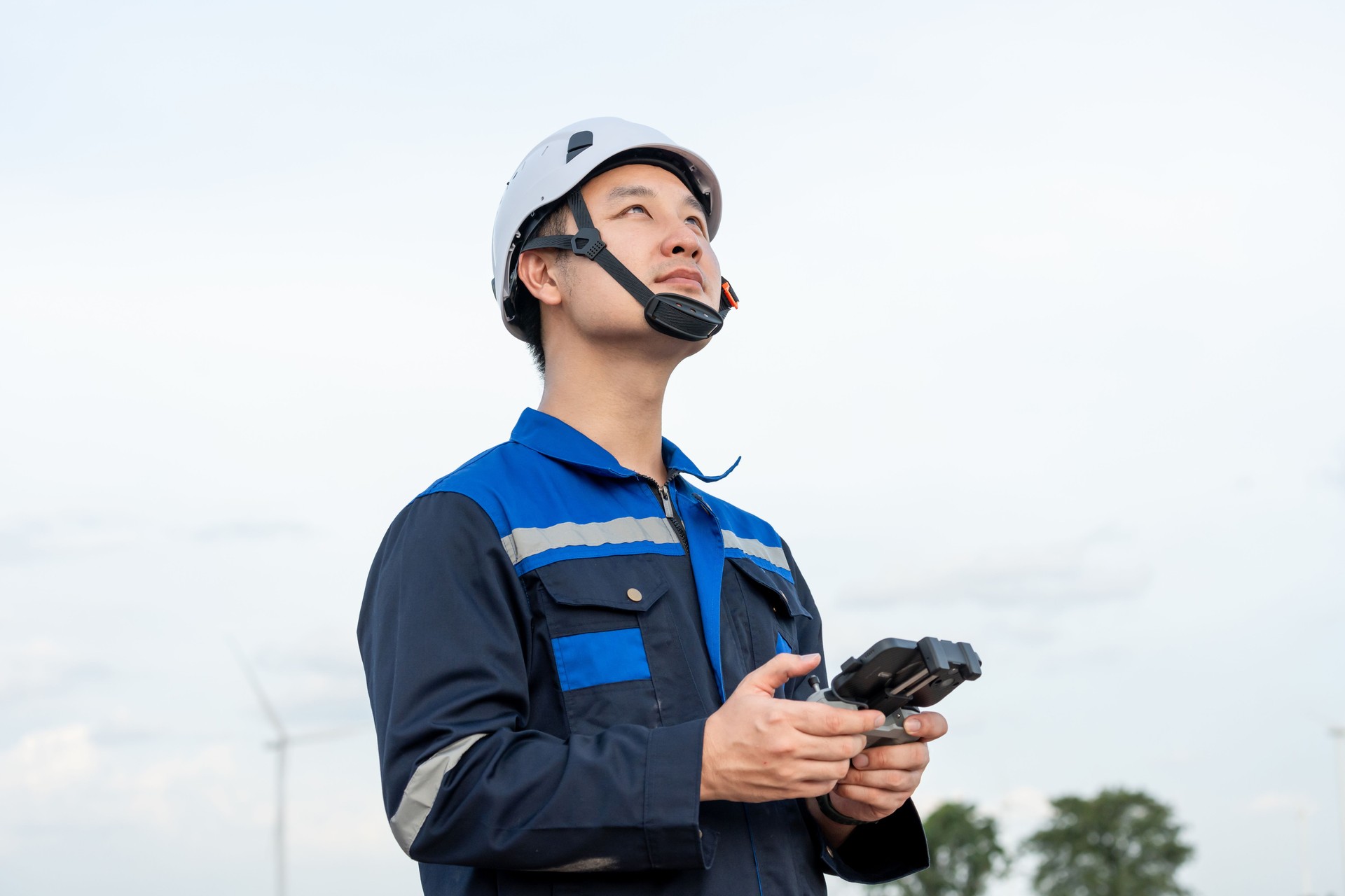 Asian male technician working outdoors in the wind turbines field. Innovation engineer men wearing safety gear, controlling flying drone work to develop environmental clean energy at windmill farm.
